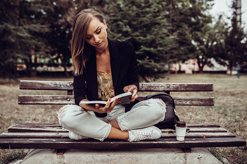 Beautiful young woman sitting on a park bench, reading a book and drinking coffee