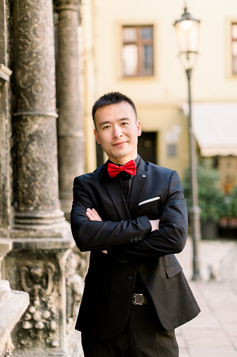 Portrait Of A Handsome Chinese Young Man Groom In Black Suit And Red Bow  Tie Looking At Camera With Arms Crossed While Standing Outdoors In Old City  Stock Photo - Download Image