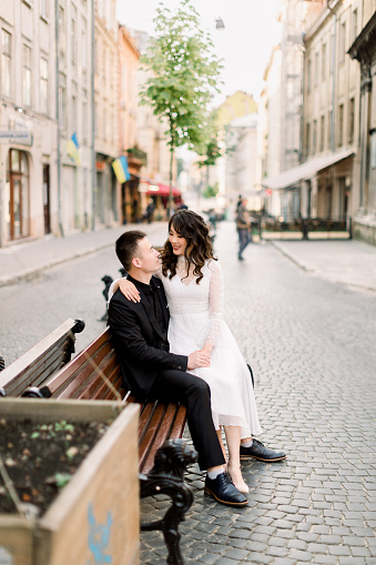 Cheerful Chinese wedding couple is holding hands while sitting on the bench on the street of old city center outdoors
