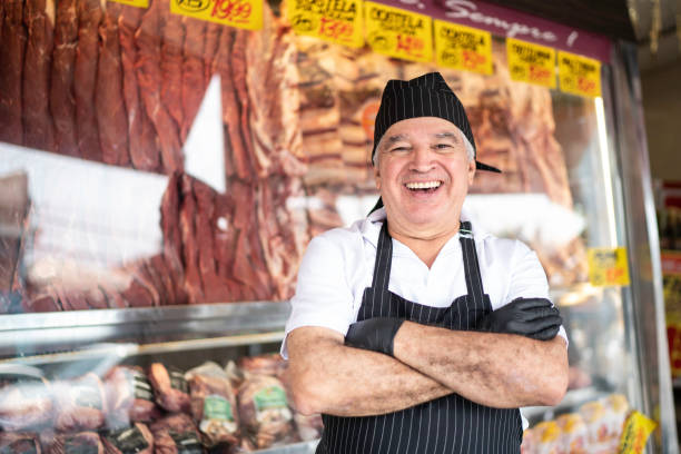 porträt eines älteren metzgers, der mit gekreuzten armen vor einer metzgerei steht - butcher meat butchers shop steak stock-fotos und bilder