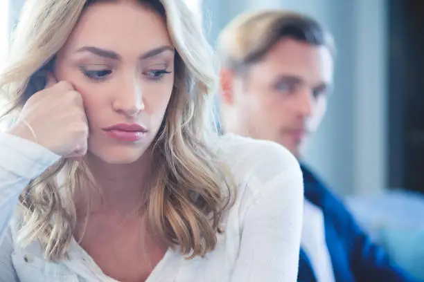 Couple fighting sitting on the sofa. The woman is close to the camera, the man is out of focus in the background. The woman looks very angry and upset. Close up