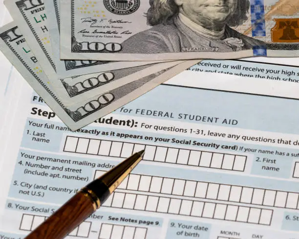 Photo of Student financial aid application forms for college tuition loans and grants with one-hundred dollar bills and ballpoint pen