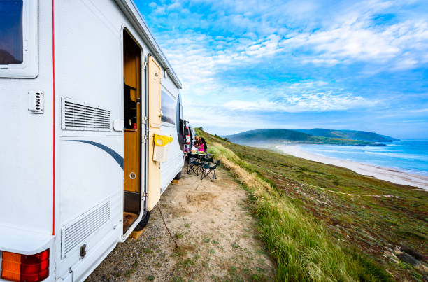 Motorhome RV and campervan are parked on a beach. Family on vacation is sitting outsides on camping chairs and table having dinner, with amazing view of the beach and ocean. Atlantic beach - Spain.