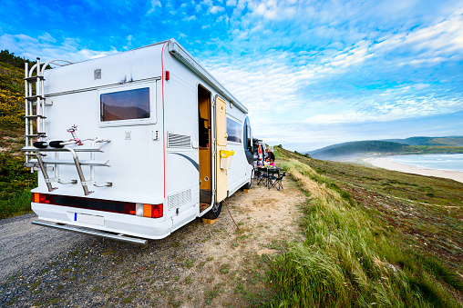 Motorhome RV and campervan are parked on a beach. Family on vacation is sitting outsides on camping chairs and table having dinner, with amazing view of the beach and ocean. Atlantic beach - Spain.