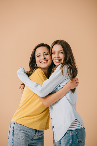 attractive and smiling friends hugging and looking at camera isolated on beige