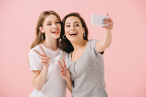 attractive and smiling women in t-shirts taking selfie and showing peace signs isolated on pink