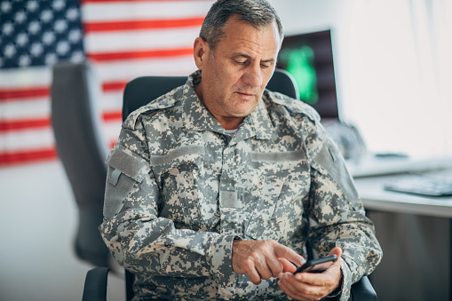 One mature man, American soldier sitting in headquarters office alone, using smart phone.
