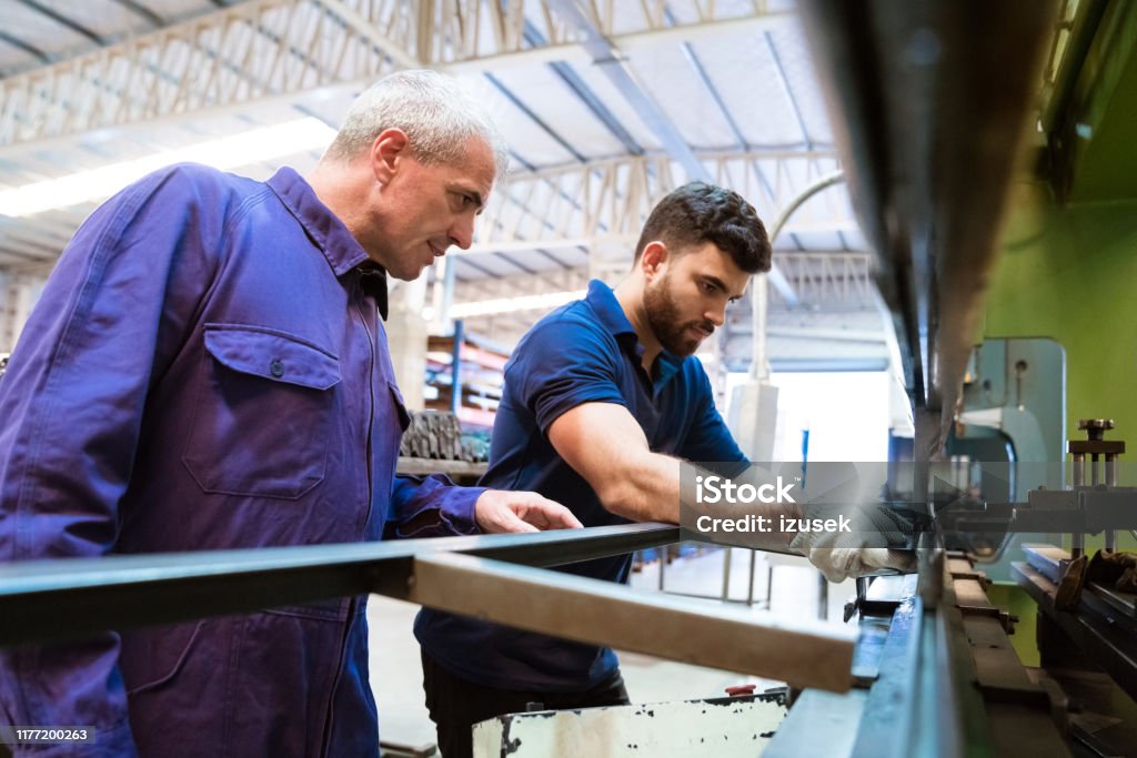 Engineers discussing over machinery in factory Mature engineer explaining apprentice over manufacturing machinery. Instructor is discussing with young trainee in factory. They are wearing uniforms. Engineer Stock Photo