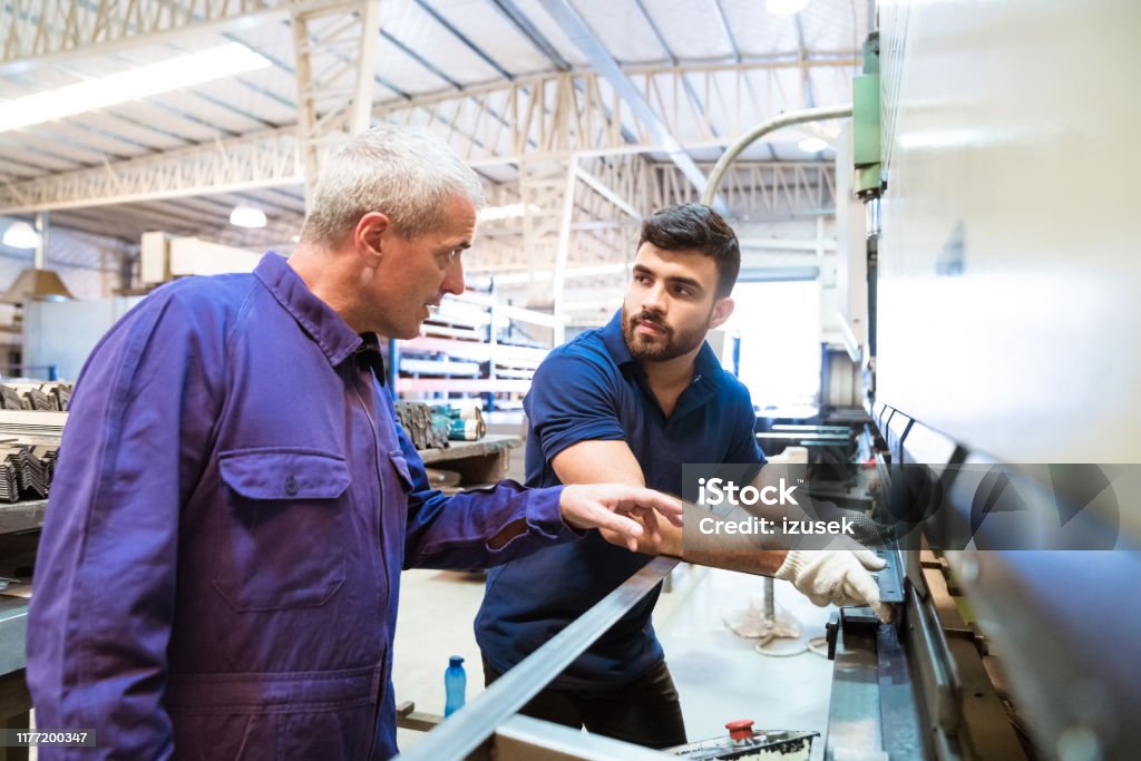 Engineers discussing over machinery in industry Male instructor explaining apprentice over manufacturing machinery. Instructor is discussing with young trainee in factory. They are wearing uniforms. Trainee Stock Photo