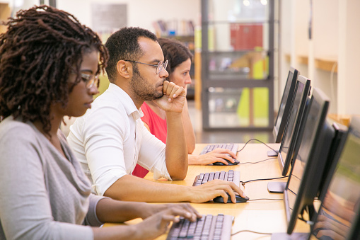 Multiracial group of students training in computer class. Line of man and women in casual sitting at table, using desktops, typing, looking at monitor. Training center concept