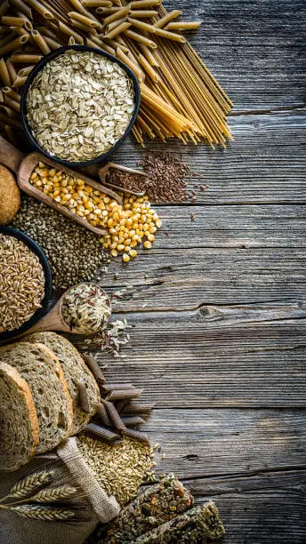 Top view of a rustic wooden table with wholegrain and cereals placed at the left border of the frame leaving useful copy space for text and/or logo at the center right. This type of food is rich of fiber and is ideal for dieting. The composition includes wholegrain sliced bread, various kinds of wholegrain pasta, wholegrain crackers, oat flakes, brown rice, spelt and flax seeds. Predominant color is brown. XXXL 42Mp studio photo taken with SONY A7rII and Zeiss Batis 40mm F2.0 CF