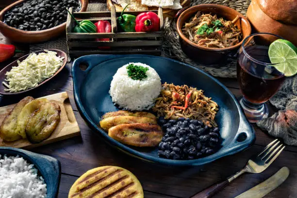 Venezuelan traditional food, Pabellon Criollo with arepas, casabe and papelon with lemon drink. Home made with white rice, black beans, fried plantains, and shredded beef. Plate on a wooden table in a rustic kitchen. Set with ingredients and shredded white cheese. Pabellon Criollo, is a main meal in the Venezuelan Culture and cuisine.