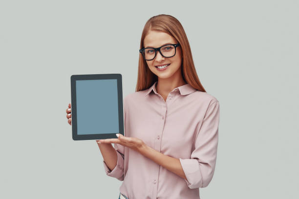 istock Attractive young woman in eyewear pointing copy space on digital tablet and smiling while standing against grey background 1177220443