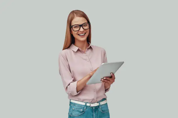 Attractive young woman in eyewear using digital tablet and smiling while standing against grey background