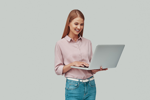 Attractive young woman using laptop and smiling while standing against grey background