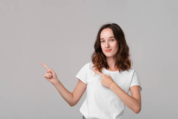 istock Pretty brunette woman in a white tee pointing at the empty space in a left side of a frame standing isolated over grey background. Copy space of advertising. 1177225140