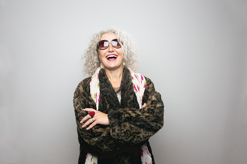 Cool mature woman,wearing sunglasses and a coat made from artificial fur, wearing scarf with floralpattern and a lot of jewellery, she is standing in front of grey background,looking into camera,smiling