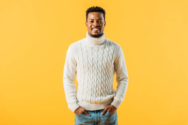 hombre afroamericano en suéter de punto que sostiene las manos en los bolsillos, sonriendo y mirando la cámara aislada en amarillo - cardigan men african ethnicity african descent fotografías e imágenes de stock