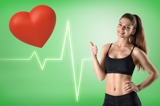Young athletic fitness girl with red heart and heart rhythm cardiogram on green background. Fitness and workout. Healthy lifestyles. People and objects.