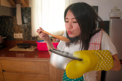 Asian home cook girl lifestyle portrait . Young happy and beautiful Chinese woman in kitchen apron and glove holding cooking pot excited and satisfied preparing soup delighted with delicious taste