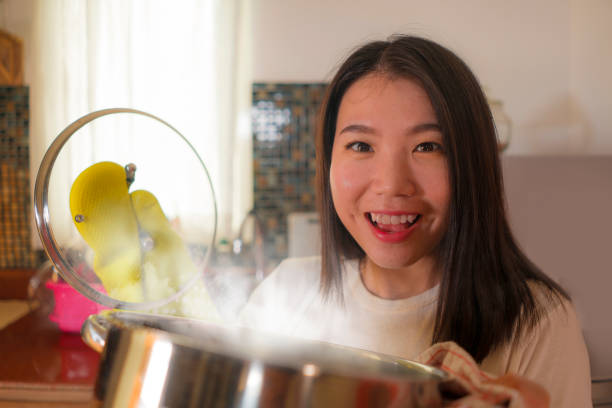 Asian home cook girl lifestyle portrait . Young happy and beautiful Korean woman in kitchen apron and glove holding cooking pot excited and satisfied preparing stew delighted with delicious aroma stock photo