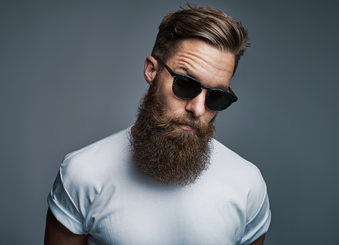 Stylish and confident young man with a long beard wearing a white t shirt and sunglasses standing against a grey background