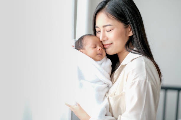 white shirt asian mother is kissing her newborn baby in bedroom in front of glass windows with white curtain to show love and family bonding - mother baby new kissing imagens e fotografias de stock