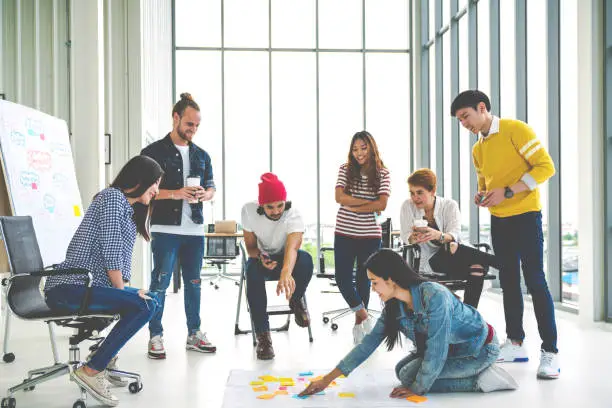 Photo of Young creative diverse group meeting and looking at project plan lay out on floor discuss or brainstorm business strategy with post note. Workshop for startup team in modern office. Happy workplace.