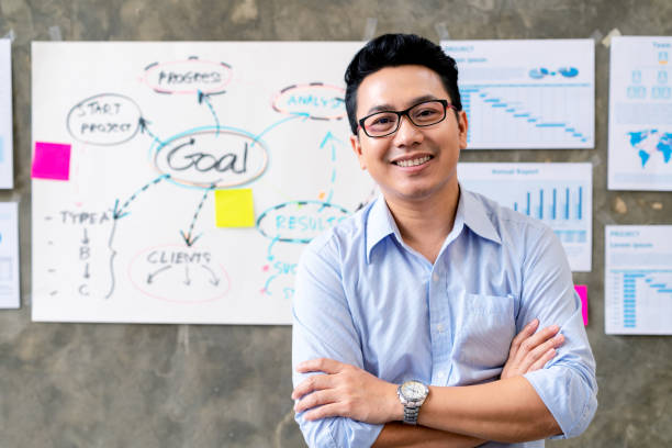 ritratto di uomo asiatico felice in camicia blu in piedi in un ambiente di lavoro smart office con piano documente e obiettivo sullo sfondo della parete. colpo alla testa di ceo o manager sorridente appoggiato tavolo con la sensazione di fiducia - crossing human arm leadership men foto e immagini stock