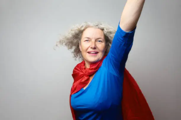 Mature woman,57 years old,wearing a blue shirt and a red cape made from a piece of fabric, her arms are raised, posing as superwoman,hair is blown away, with the help of a fan ,as if flying. The woman has beautiful long, grey and curly hair