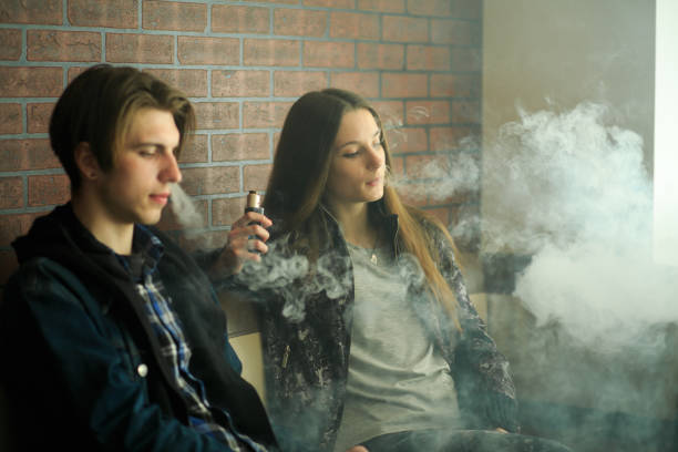 vapeティーンエイジャー。サングラスと若いハンサムな男の若いかわいい女の子は、vapeバーで電子タバコを吸います。健康に有害な悪い習慣。 - 電子タバコ ストックフォトと画像