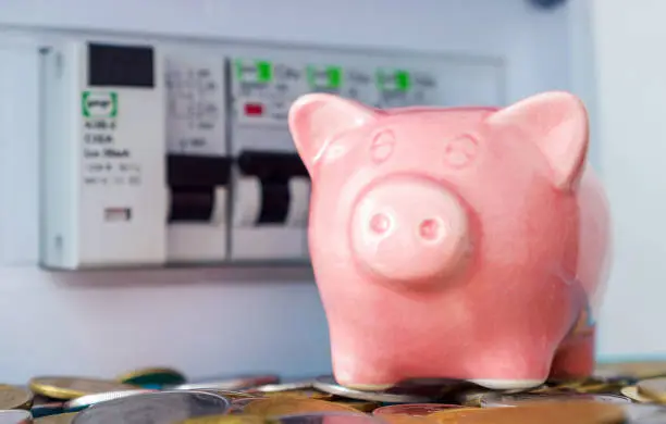 Photo of Pink piggy bank with coins near a residential electrical panel. The symbolic image of the need to save electricity, pay for electricity consumed, energy efficiency apartment building.