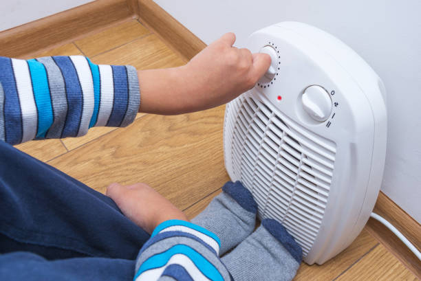A young boy warms himself near an electric fan heater, sitting on the floor at home. Part of body, selective focus. stock photo