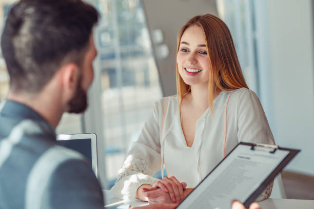 uomo d'affari gioioso e di successo che pone domande ai candidati, seduto alla scrivania sul posto di lavoro sulla sedia - recruiter foto e immagini stock