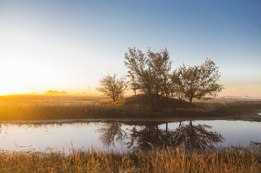 A small pond reflects trees and the rising sun on a foggy morning in rural Kansas.