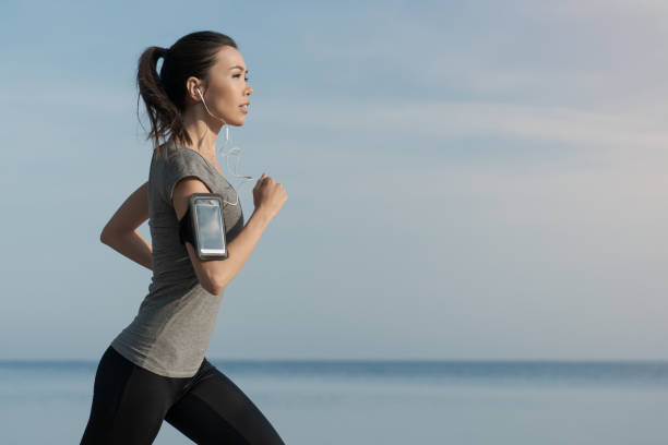 outdoor shot of young sports asian woman running along the sea shore in morning - asian ethnicity jogging female women imagens e fotografias de stock