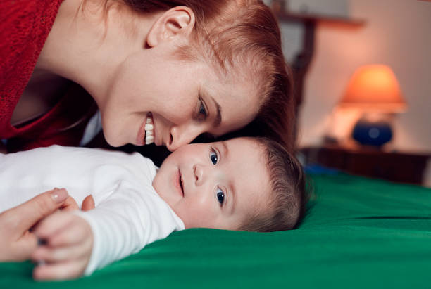 Little baby girl lying on her back on green bed, wearing Christmas hat; father kissing her