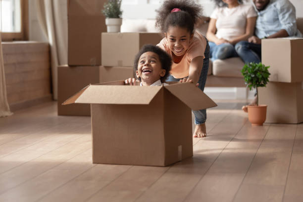 Sister riding younger brother who is sitting in cardboard box African older sister riding younger cute brother who is sitting in cardboard box, on background big carton boxes stuff and parents sitting on couch enjoy new modern house resting at moving day concept sibling stock pictures, royalty-free photos & images