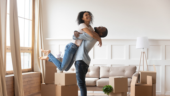 African husband lifting up beloved wife happy family celebrating relocation day full length view, stack of carton boxes on background, moving at new home, property owners, lease loan mortgage concept