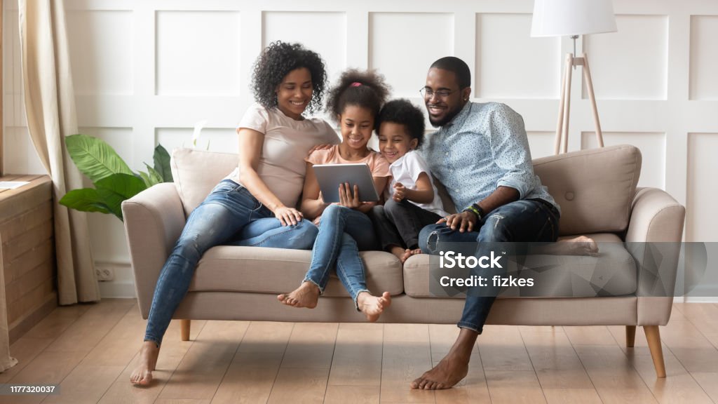 African full family using tablet computer resting on couch Concept of parental control, modern wireless technology usage, leisure activities at home with children, african full family with daughter and son resting on sofa use tablet computer having fun online Family Stock Photo