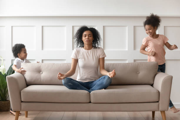 mentre i bambini rumorosi che corrono giovane madre africana medita sul divano - spirituality yoga zen like meditating foto e immagini stock