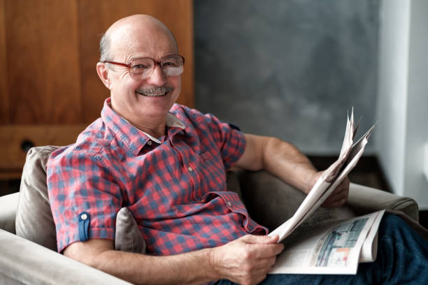 Handsome mature hispanic man sitting in living room, reading newspaper, smiling.