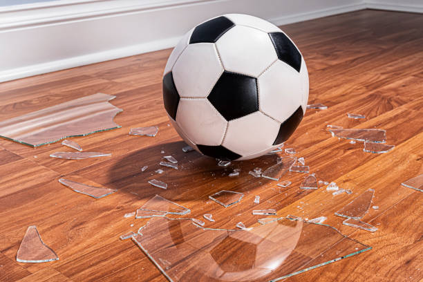 футбольный мяч с разбитым стеклом из окна на полу - shattered glass broken window damaged стоковые фото и изображения