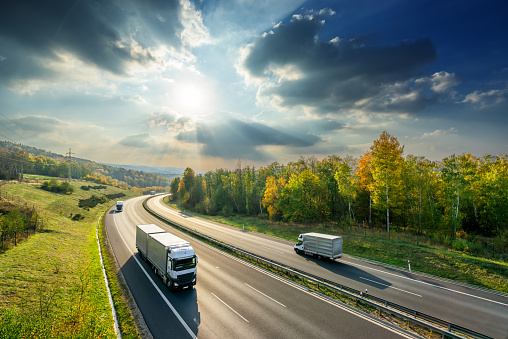 Camiones y furgoneta de reparto que conducen por la carretera del asfalto entre bosque caducifolio en colores otoñales bajo el sol radiante y nubes dramáticas. Vista desde arriba. photo