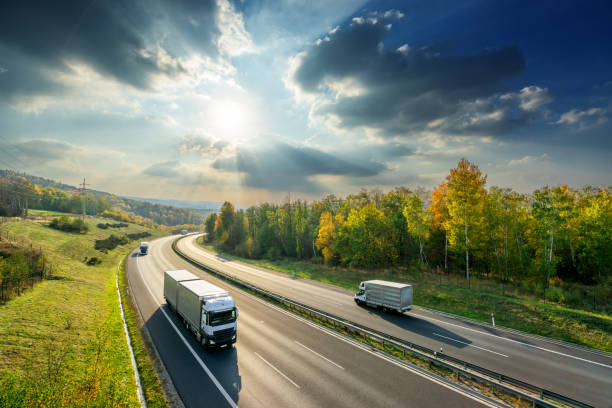 輝く太陽と劇的な雲の下で秋の色の落葉林の間のアスファルトハイウェイを運転するトラックと配達バン。上から見る。 - truck ストックフォトと画像