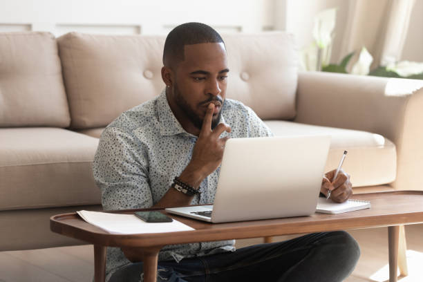 Young african american guy studying on online courses. Concentrated young african american black guy sitting on heated floor at modern coffee table in living room, looking at laptop screen, online courses studying, listening to coach, writing down notes. curiosity stock pictures, royalty-free photos & images