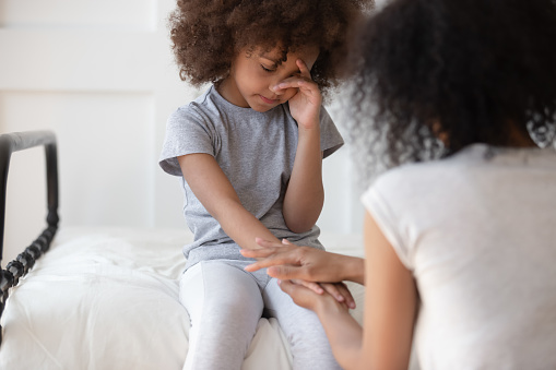 Mixed race mommy holding hand of upset little cute girl sitting on bed, wiping tears. African american worried woman talking, supporting, comforting small adopted daughter, suffering from bullying.