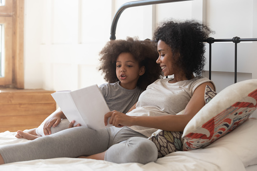 Happy smiling single mixed race mother lying on bed with adorable daughter, holding book, reading fairytale, teaching cute kid to read, enjoying morning weekend time in comfortable bedroom together.