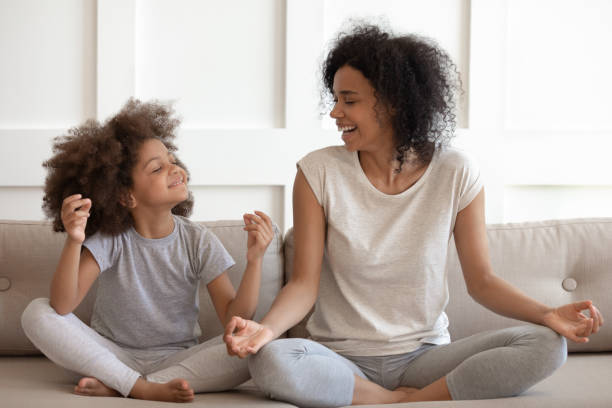 Excited african american woman practicing yoga with little daughter. Excited african american woman teaching cute little adorable daughter meditating and practicing yoga exercises. Happy mixed race family sitting on comfy couch in lotus position, having fun together. mindfulness kids stock pictures, royalty-free photos & images