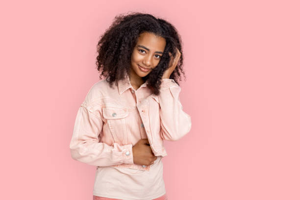 kür. afrikanische mädchen in jeansjacke stehen isoliert auf rosa posiert berührende haare lächelnd niedlich - afro amerikanischer herkunft stock-fotos und bilder